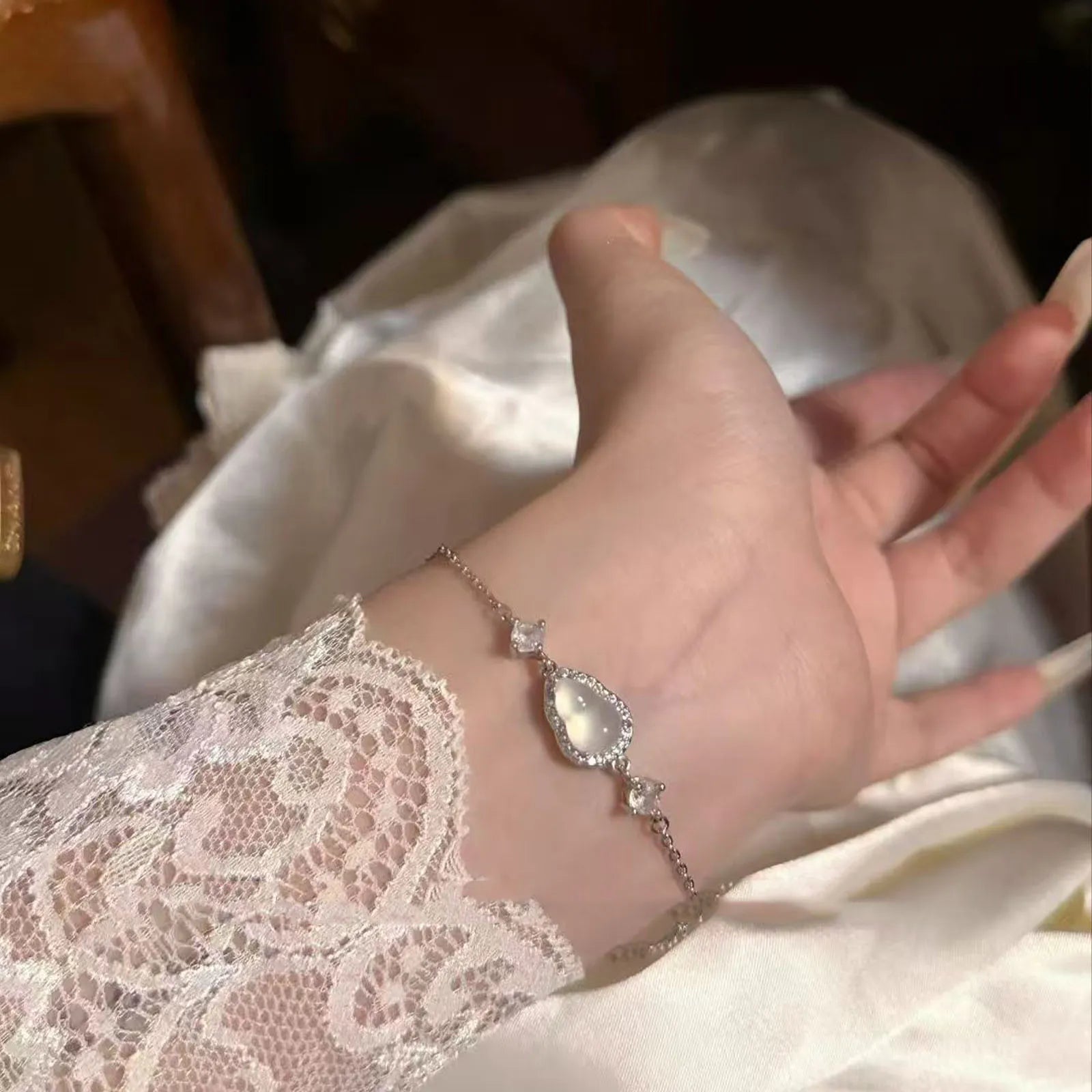 A delicate silver-toned bracelet featuring a teardrop-shaped luminous gem (resembling moonstone) and sparkling accents, worn on a wrist with lace detailing against soft satin fabric — ideal as a complimentary gift.